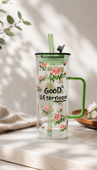 Alt Text: A tall, clear glass mug adorned with a pink floral pattern and the text "Good Afternoon" printed in black. The mug features a translucent green handle, lid, and glass straw, displayed on a light-colored countertop under soft, sunny daylight shadows.