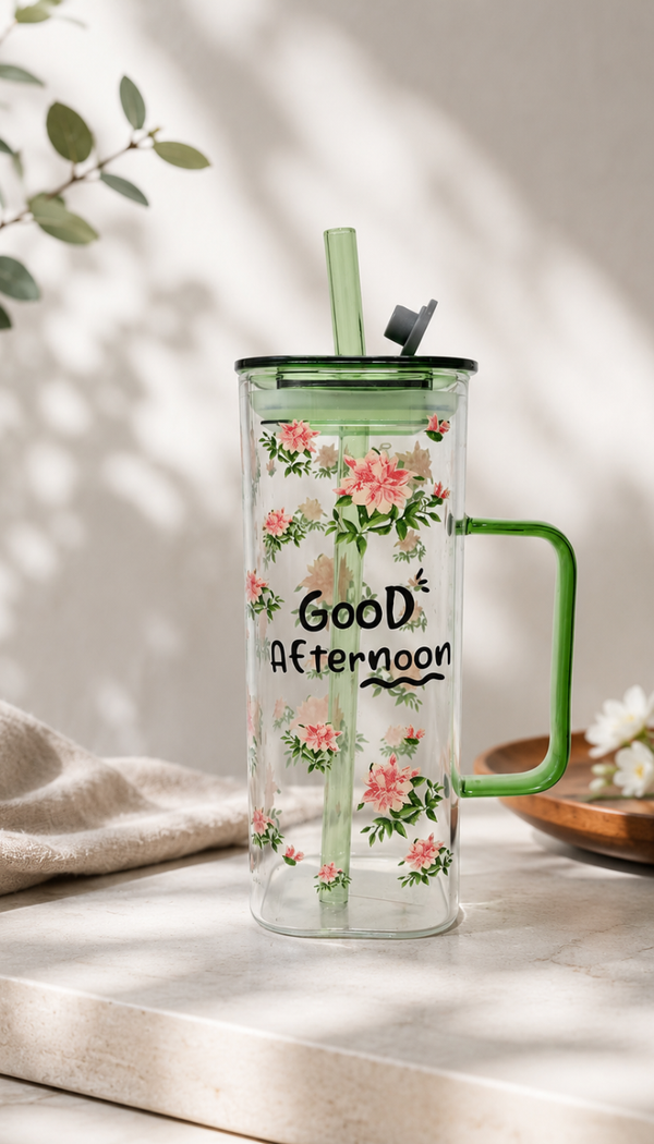 Alt Text: A tall, clear glass mug adorned with a pink floral pattern and the text "Good Afternoon" printed in black. The mug features a translucent green handle, lid, and glass straw, displayed on a light-colored countertop under soft, sunny daylight shadows.