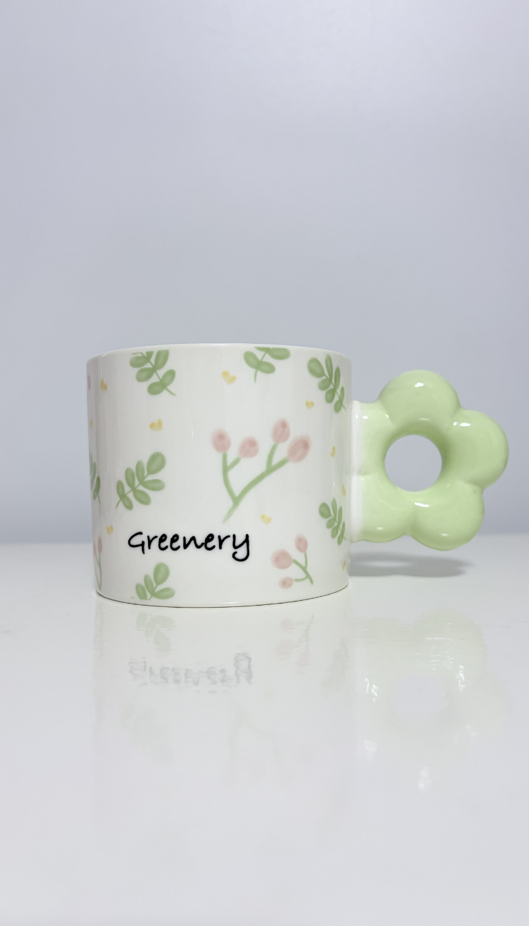 Alt Text: A cylindrical white ceramic mug decorated with a delicate pattern of green leaves, tiny pink flower buds, and the word "Greenery" printed in a handwritten font. The mug features a unique chunky handle shaped like a pastel green flower, sitting on a glossy white surface with a soft reflection against a plain background.