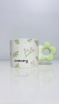 Alt Text: A cylindrical white ceramic mug decorated with a delicate pattern of green leaves, tiny pink flower buds, and the word "Greenery" printed in a handwritten font. The mug features a unique chunky handle shaped like a pastel green flower, sitting on a glossy white surface with a soft reflection against a plain background.