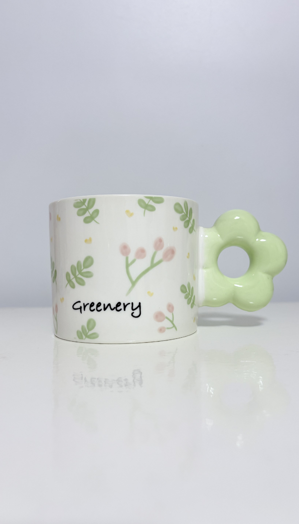 Alt Text: A cylindrical white ceramic mug decorated with a delicate pattern of green leaves, tiny pink flower buds, and the word "Greenery" printed in a handwritten font. The mug features a unique chunky handle shaped like a pastel green flower, sitting on a glossy white surface with a soft reflection against a plain background.