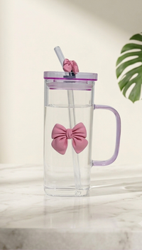 Alt Text: An elegant, rectangular clear glass mug filled with water, equipped with a purple lid and a transparent glass straw. The cup is adorned with a prominent pastel pink bow accent in the center and a small matching bow accessory on the straw, shot on a bright marble tabletop with a clean, minimalist background.