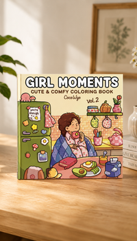 A cozy product mockup of a coloring book titled "Girl Moments Cute & Comfy Coloring Book Vol. 2" displayed on a wooden table. The cover features a charming cartoon illustration of a girl wrapped in a blue blanket eating a donut in her kitchen, next to a green refrigerator with magnets, and a table with breakfast, toast, and avocado. A green houseplant and ceramic vases are in the sunlit background.
